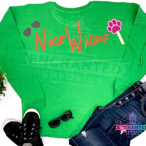 May include: Green long-sleeved shirt with the text "Nick Wilde" in orange glitter. The shirt has a paw print lollipop on the right sleeve and sunglasses on the left sleeve.