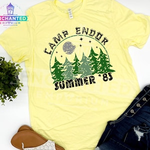 May include: A light yellow t-shirt with a graphic that reads "CAMP ENDOR SUMMER '83". The design features a forest scene with a moon and stars. The shirt is paired with blue jeans and sandals.