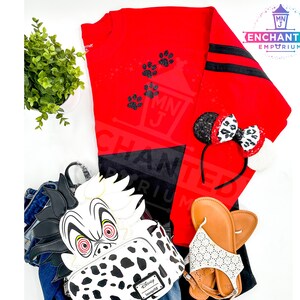 K&ouml;nnte beinhalten: Ein rot-schwarzes Langarmshirt mit schwarzen Pfotenabdr&uuml;cken. Ein schwarz-wei&szlig;er Dalmatiner-Rucksack mit roten Augen. Ein schwarzes Stirnband mit einer rot-wei&szlig;en Schleife. Ein Paar wei&szlig;e Sandalen mit schwarzen Riemen. Ein Paar braune Sandalen.