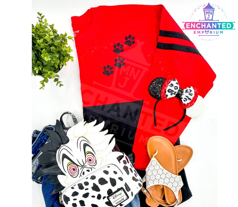 K&ouml;nnte beinhalten: Ein rot-schwarzes Langarmshirt mit schwarzen Pfotenabdr&uuml;cken. Ein schwarz-wei&szlig;er Dalmatiner-Rucksack mit roten Augen. Ein schwarzes Stirnband mit einer rot-wei&szlig;en Schleife. Ein Paar wei&szlig;e Sandalen mit schwarzen Riemen. Ein Paar braune Sandalen.