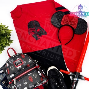 May include: Red and black long-sleeved shirt with a black Darth Vader graphic. The shirt has the text "ENCHANTED EMPORIUM" on it.  A black and red mini backpack with a Star Wars theme. A black and red headband with black glitter Minnie Mouse ears. A red lightsaber.