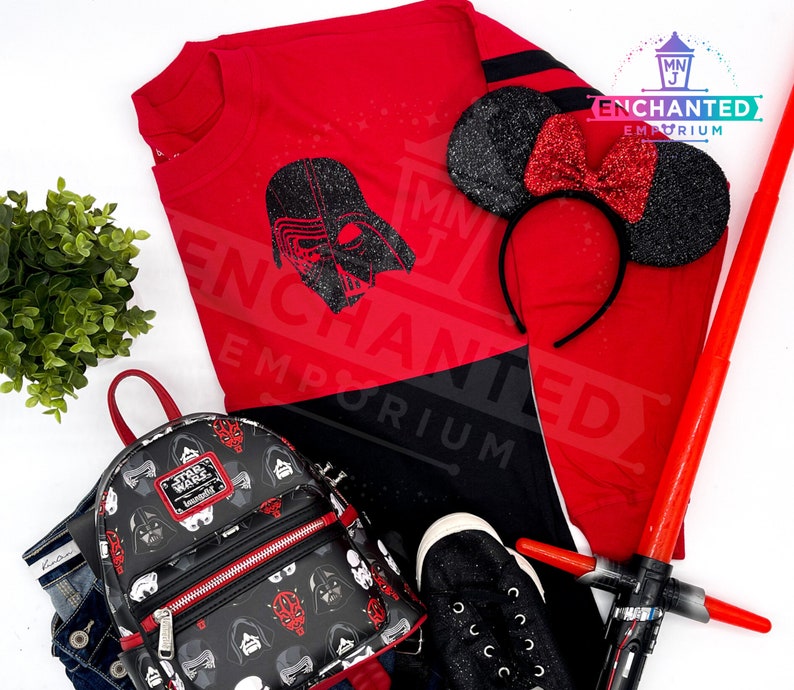 May include: Red and black long-sleeved shirt with a black Darth Vader graphic. The shirt has the text "ENCHANTED EMPORIUM" on it.  A black and red mini backpack with a Star Wars theme. A black and red headband with black glitter Minnie Mouse ears. A red lightsaber.