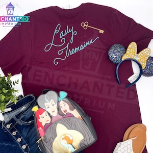 May include: Burgundy t-shirt with the text "Lady Tremaine" in blue script. The shirt is paired with a black backpack featuring the faces of Cinderella's step-family, a pair of blue and gold glitter Minnie Mouse ears, and a pair of brown sandals.