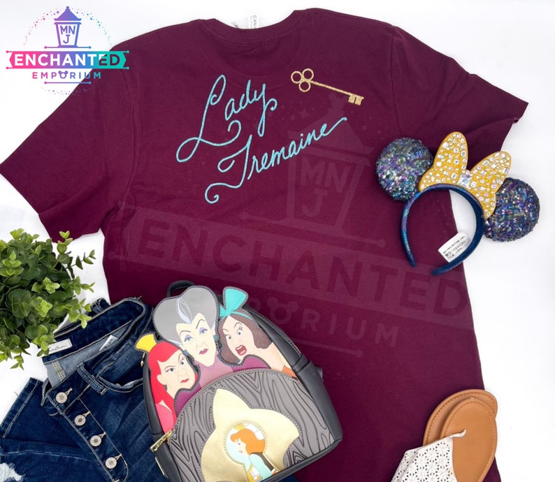 May include: Burgundy t-shirt with the text "Lady Tremaine" in blue script. The shirt is paired with a black backpack featuring the faces of Cinderella's step-family, a pair of blue and gold glitter Minnie Mouse ears, and a pair of brown sandals.