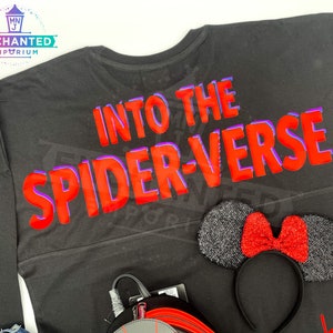May include: Black long-sleeved shirt with the text "Into the Spider-Verse" in red with a purple outline. A black headband with a red glitter bow is in the bottom right corner.