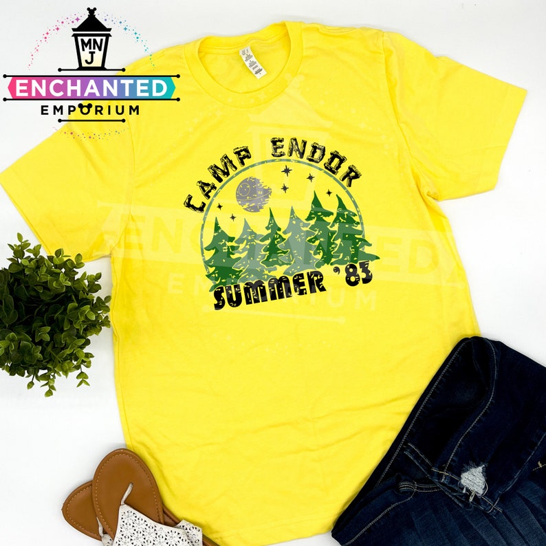 May include: Yellow t-shirt with a graphic that reads "Camp Endor Summer '83". The design includes a moon, stars, and green trees. The shirt is paired with blue denim jeans and sandals.