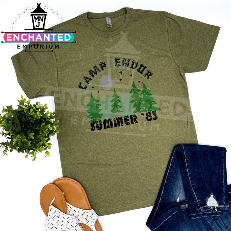 May include: Olive green t-shirt with the text "Camp Endor Summer '83" in black and green. The design includes a forest scene with trees and a moon. The shirt is paired with blue jeans and sandals.