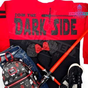 May include: Red and black long-sleeved shirt with the text "Join the Dark Side" in black glitter. The shirt is paired with a black and red Minnie Mouse headband, a black backpack with Star Wars characters, a red lightsaber, and black sneakers.