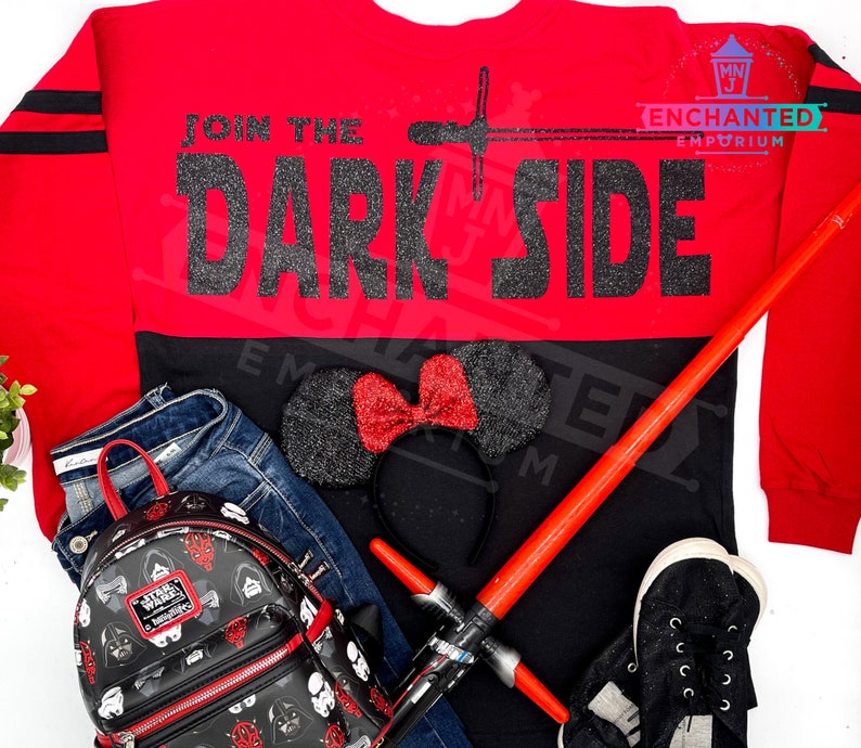 May include: Red and black long-sleeved shirt with the text "Join the Dark Side" in black glitter. The shirt is paired with a black and red Minnie Mouse headband, a black backpack with Star Wars characters, a red lightsaber, and black sneakers.