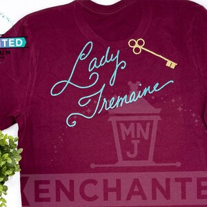 May include: Burgundy t-shirt with a teal blue glitter design that says "Lady Tremaine" with a gold key above the text. The shirt has a small logo on the bottom right corner that says "Enchanted".