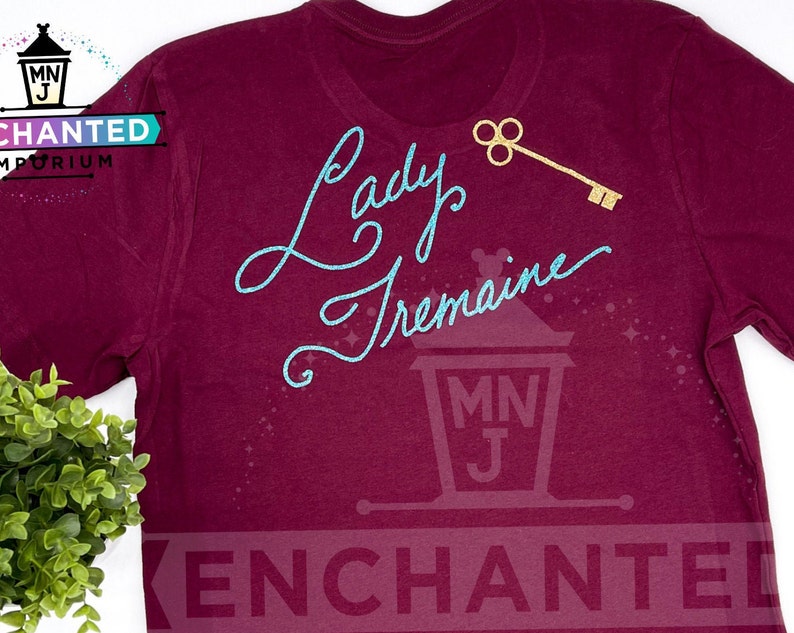 May include: Burgundy t-shirt with a teal blue glitter design that says "Lady Tremaine" with a gold key above the text. The shirt has a small logo on the bottom right corner that says "Enchanted".