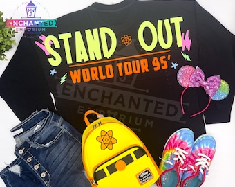 May include: Black sweatshirt with the text "Stand Out World Tour 95'" in neon yellow and orange. The sweatshirt is paired with a yellow backpack with an atom symbol, tie-dye trainers, and pink glitter Minnie Mouse ears.