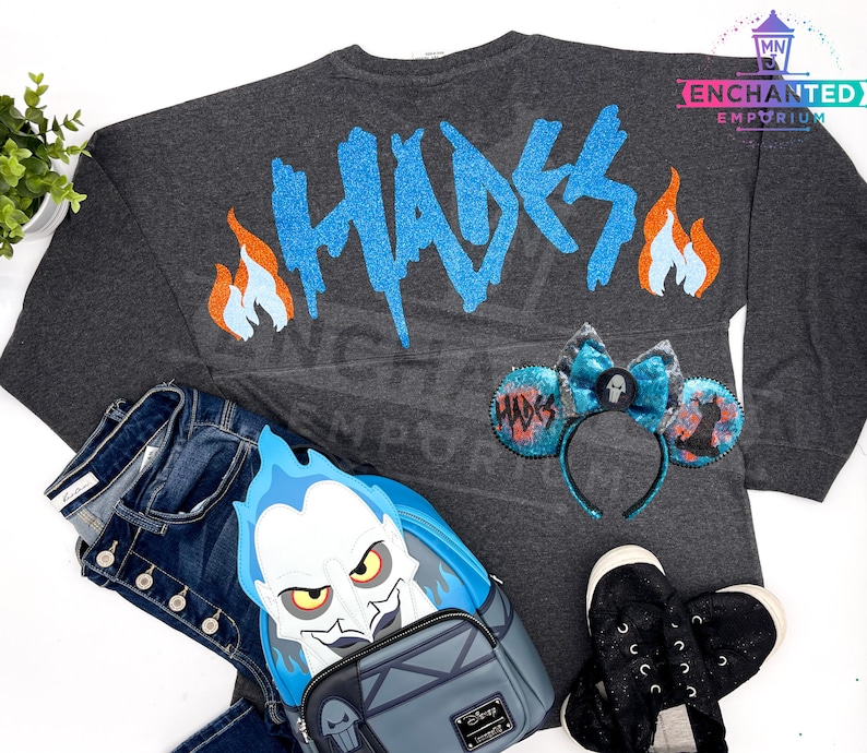 May include: A gray long-sleeved shirt with the word "Hades" in blue glitter on the back. The shirt is paired with a blue and black Hades-themed backpack, a pair of black shoes, and a pair of blue and black mouse ears with a skull and crossbones design.