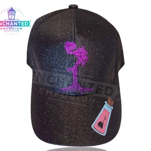 Op de afbeelding: Zwarte glitter trucker cap met een paars glitter ontwerp van een silhouet van een persoon die een fles vasthoudt. De fles heeft een roze vloeistof en een roze silhouet van een wezen erin.