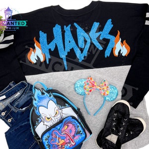 May include: A black long-sleeve shirt with a grey and black color block design. The shirt has the word "Hades" written in blue glitter on the back with orange flames on either side. The shirt is paired with a black backpack with a blue and white glitter design of Hades from Disney's Hercules. The backpack also features a blue and purple glitter design of the three-headed dog Cerberus. The shirt is also paired with a blue headband with a pink and white polka dot bow and black glitter sneakers.
