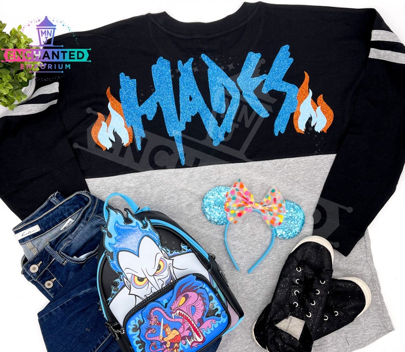 May include: A black long-sleeve shirt with a grey and black color block design. The shirt has the word "Hades" written in blue glitter on the back with orange flames on either side. The shirt is paired with a black backpack with a blue and white glitter design of Hades from Disney's Hercules. The backpack also features a blue and purple glitter design of the three-headed dog Cerberus. The shirt is also paired with a blue headband with a pink and white polka dot bow and black glitter sneakers.