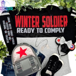 May include: A black and gray long-sleeved shirt with the text "WINTER SOLDIER READY TO COMPLY" in red glitter. The shirt has a gray and black color block design. The shirt is paired with a gray backpack with a red star, a silver chain necklace with a dog tag, a pair of black sneakers, and a black headband with a silver bow and a red, white, and blue star.