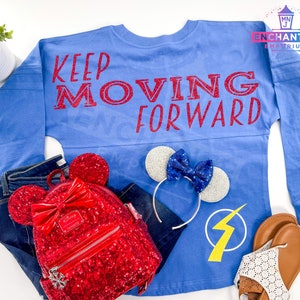 May include: A blue long-sleeved shirt with the text "Keep Moving Forward" in red glitter. The shirt is laid out on a white surface with a red sequined Minnie Mouse backpack, a silver headband with blue bow, and brown sandals.