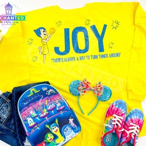 May include: Yellow long-sleeved shirt with a blue glitter "JOY" graphic and a cartoon character holding a yellow ball. The shirt has the text "There's always a way to turn things around".