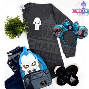 May include: A gray sweatshirt with a white skull graphic and the text "ENCHANTED EMPORIUM" printed on it. A blue and black headband with a skull graphic and the text "HADES" is also pictured. A blue and gray backpack with a Hades character graphic is also pictured.