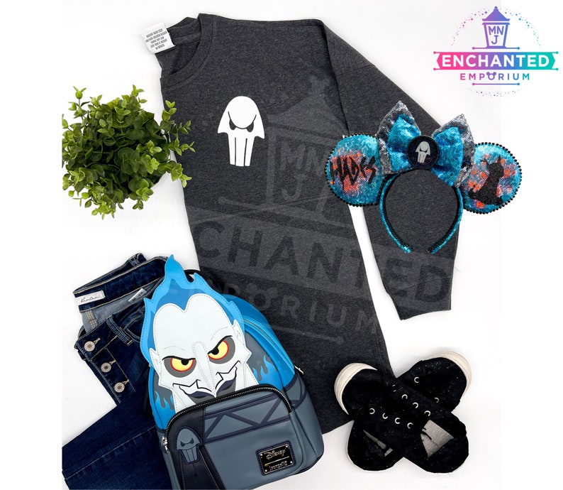 May include: A gray sweatshirt with a white skull graphic and the text "ENCHANTED EMPORIUM" printed on it. A blue and black headband with a skull graphic and the text "HADES" is also pictured. A blue and gray backpack with a Hades character graphic is also pictured.