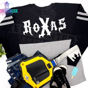 May include: A black and gray long-sleeved shirt with white lettering that says "ROXAS" on the back. The shirt has a gray stripe on the sleeves. A yellow and black key-shaped bag is in the bottom left corner of the image.