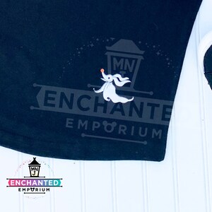 May include: Black t-shirt featuring a white ghost character holding a small orange ball. The text "ENCHANTED EMPORIUM" is in white, along with the text "MNJ" in a small white circle above the ghost.