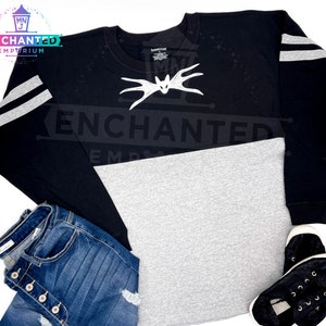 May include: Black and gray long-sleeved shirt with white stripes on the sleeves. The shirt has a gray bottom half and a black top half. The shirt has a white bat design on the front with the word "ENCHANTED" printed on the shirt.