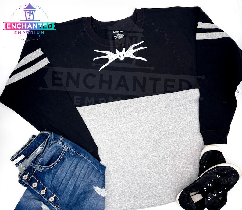 May include: Black and gray long-sleeved shirt with white stripes on the sleeves. The shirt has a gray bottom half and a black top half. The shirt has a white bat design on the front with the word "ENCHANTED" printed on the shirt.