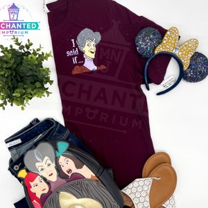 May include: A maroon t-shirt with a glittery graphic of a woman with the text "I said if..." and the words "Enchanted Emporium" printed on the bottom. The shirt is paired with a pair of brown sandals, a black and grey backpack with a glittery graphic of three women, and a pair of sparkly Minnie Mouse ears.