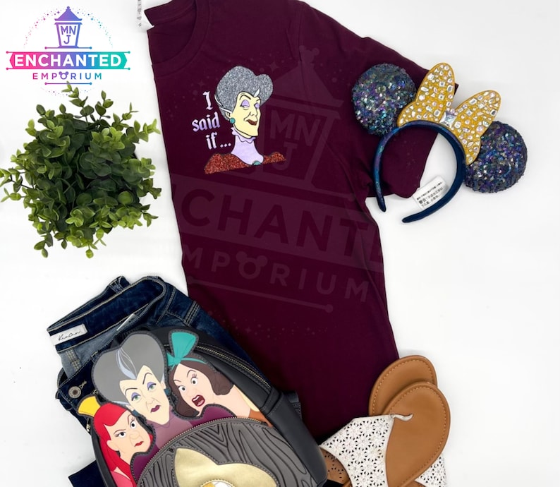 May include: A maroon t-shirt with a glittery graphic of a woman with the text "I said if..." and the words "Enchanted Emporium" printed on the bottom. The shirt is paired with a pair of brown sandals, a black and grey backpack with a glittery graphic of three women, and a pair of sparkly Minnie Mouse ears.