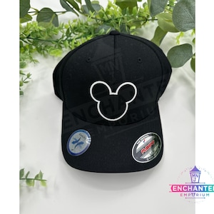 May include: Black baseball cap with a white Mickey Mouse silhouette embroidered on the front. The cap has a black brim and a black adjustable strap with a plastic buckle. The cap has a white "Duckbill" logo on the side and a black "Flexfit" logo on the back.