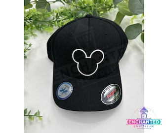 May include: Black baseball cap with a white Mickey Mouse silhouette embroidered on the front. The cap has a black brim and a black adjustable strap with a plastic buckle. The cap has a white "Duckbill" logo on the side and a black "Flexfit" logo on the back.