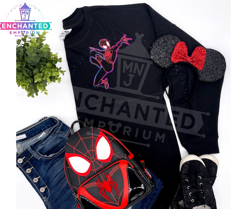 May include: Black long-sleeved shirt featuring a neon-coloured graphic of Spider-Man. The shirt is paired with a red bow headband and a black backpack with a Spider-Man design.