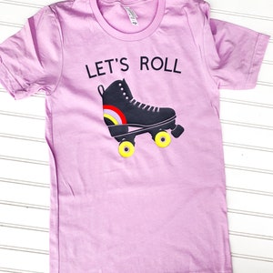 Peut inclure: Un t-shirt rose p&acirc;le avec le texte "Let's Roll" et un patin &agrave; roulettes noir avec des roues jaunes.