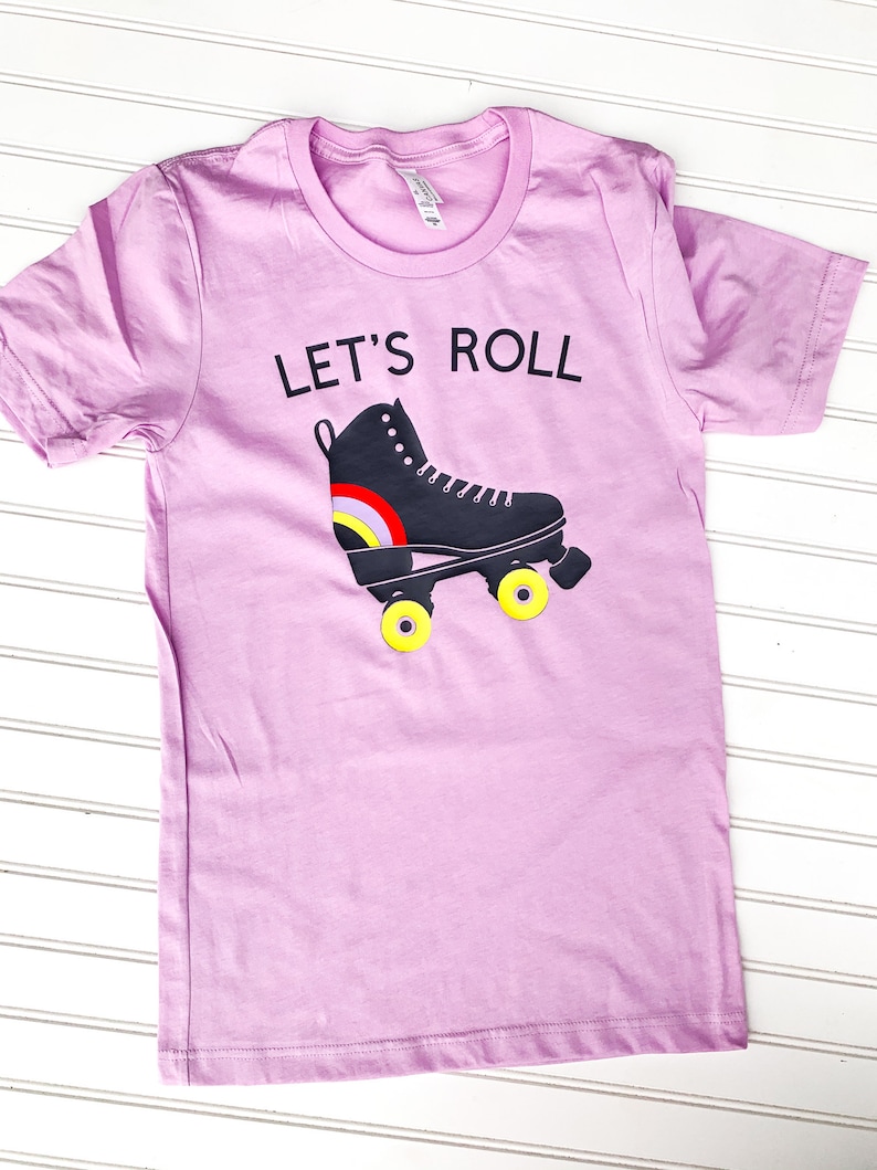 Peut inclure: Un t-shirt rose p&acirc;le avec le texte "Let's Roll" et un patin &agrave; roulettes noir avec des roues jaunes.