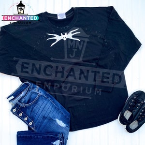 May include: Black long-sleeved shirt with a white graphic of a bat and the text "MNJ ENCHANTED EMPORIUM". The shirt is paired with blue jeans and black shoes.