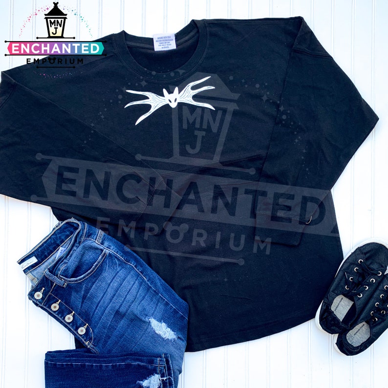 May include: Black long-sleeved shirt with a white graphic of a bat and the text "MNJ ENCHANTED EMPORIUM". The shirt is paired with blue jeans and black shoes.