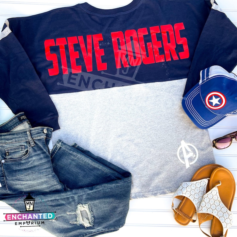 Puede incluir: Una camiseta de manga larga azul marino y gris con el texto "STEVE ROGERS" en letras rojas. La camiseta se combina con jeans azules, una gorra de b&eacute;isbol azul con una estrella blanca y sandalias marrones.