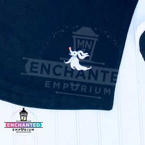May include: Black t-shirt with a white ghost character and the text "ENCHANTED EMPORIUM" in white. The ghost is holding a small orange ball. The text "MNJ" is in a small white circle above the ghost.