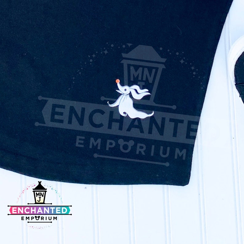 May include: Black t-shirt with a white ghost character and the text "ENCHANTED EMPORIUM" in white. The ghost is holding a small orange ball. The text "MNJ" is in a small white circle above the ghost.