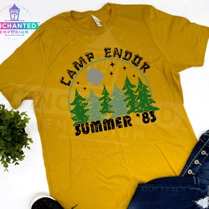 May include: Mustard yellow t-shirt with a graphic design. The design features the text "CAMP ENDOR SUMMER '83" in black, with a forest scene in green and blue. The shirt is a casual wear item.
