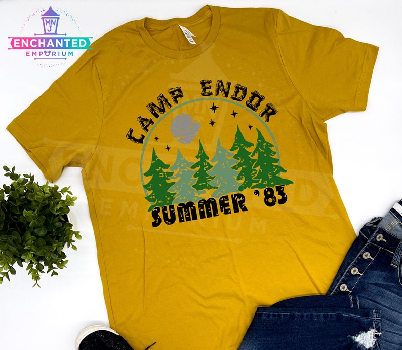 May include: Mustard yellow t-shirt with a graphic design. The design features the text "CAMP ENDOR SUMMER '83" in black, with a forest scene in green and blue. The shirt is a casual wear item.