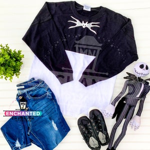 May include: A black and white long-sleeved shirt with a white bat design on the front. The shirt is a baseball style with black sleeves and a white body. The shirt is laying on a white surface with a pair of black shoes, blue jeans, and a Jack Skellington doll.