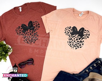 May include: Two t-shirts with a black and brown leopard print Minnie Mouse head design. The shirt on the left is a rust color and the shirt on the right is a peach color.