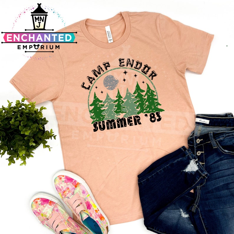 May include: Peach-colored t-shirt with the text "Camp Endor Summer '83" in a circular design featuring trees, a moon, and stars. The shirt is paired with dark blue jeans and colorful slip-on sneakers.
