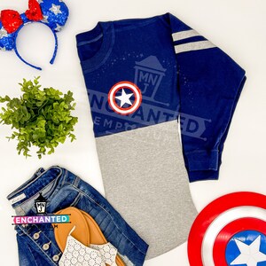 Puede incluir: Una camiseta de manga larga azul marino con un dise&ntilde;o de bloques de color gris y blanco. La camiseta presenta un logotipo de Capit&aacute;n Am&eacute;rica rojo, blanco y azul en el pecho. La camiseta tiene rayas blancas en las mangas.