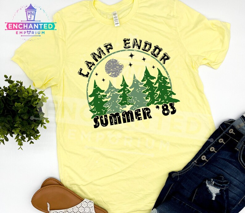 May include: A light yellow t-shirt with a graphic that reads "CAMP ENDOR SUMMER '83". The design features a forest scene with a moon and stars. The shirt is paired with blue jeans and sandals.