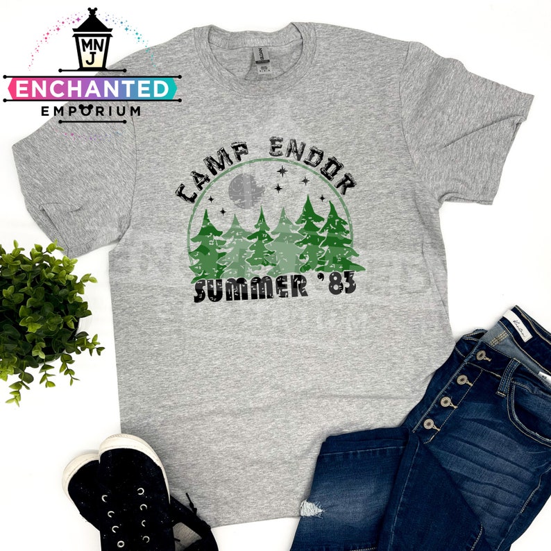 May include: A heather gray t-shirt with the text "Camp Endor Summer '83" in a distressed green and black design. The graphic includes a forest silhouette and a moon. The shirt is paired with blue jeans and black sneakers.