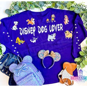 May include: A purple "Disney Dog Lover" spirit jersey with cartoon dog graphics. Accessories include a sequined backpack, Minnie Mouse ears, and sandals. The image is styled with denim and greenery.